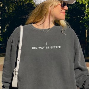 May include: A dark gray sweatshirt with the text "HIS WAY IS BETTER" and a cross graphic. The sweatshirt is worn with a white baseball cap and sunglasses. A coffee cup is held in the hand. The Red Prairie Apparel logo is visible.
