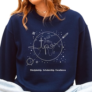 May include: Navy blue sweatshirt with a white graphic of a globe, cross, and pencil. The text "TIPS" is in the center of the globe, with the words "Discipleship. Scholarship. Excellence" below. The sweatshirt is a crew neck.