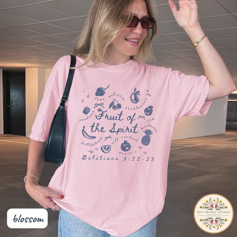 May include: A light pink t-shirt with the text "Fruit of the Spirit" and related illustrations in navy blue. The shirt also includes the Bible verse Galatians 5:22-23. The word "blossom" is in the bottom left corner.
