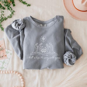 May include: A gray sweatshirt with a white graphic of a nativity scene, including a star and the text "a thrill of hope, the weary world rejoices." The sleeves are rolled up. The sweatshirt is on a cream-colored surface.