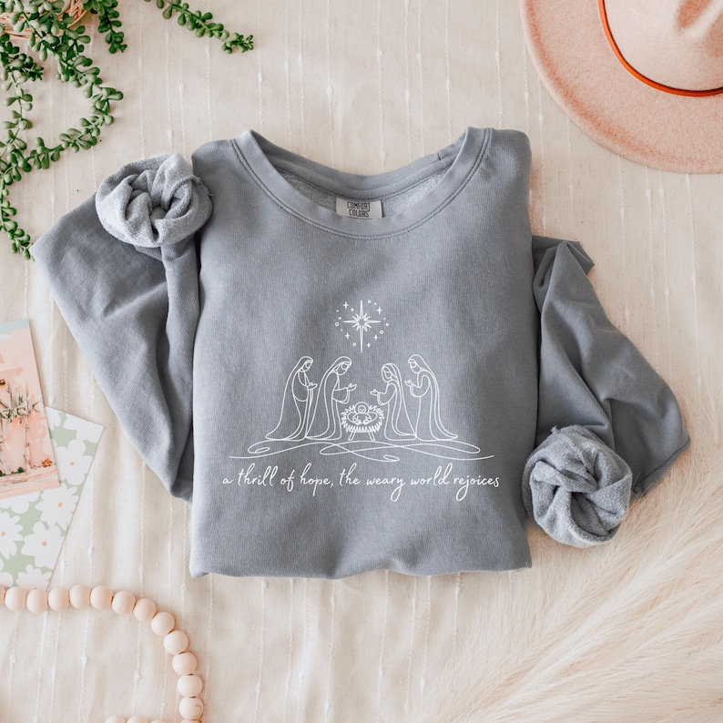 May include: A gray sweatshirt with a white graphic of a nativity scene, including a star and the text "a thrill of hope, the weary world rejoices." The sleeves are rolled up. The sweatshirt is on a cream-colored surface.