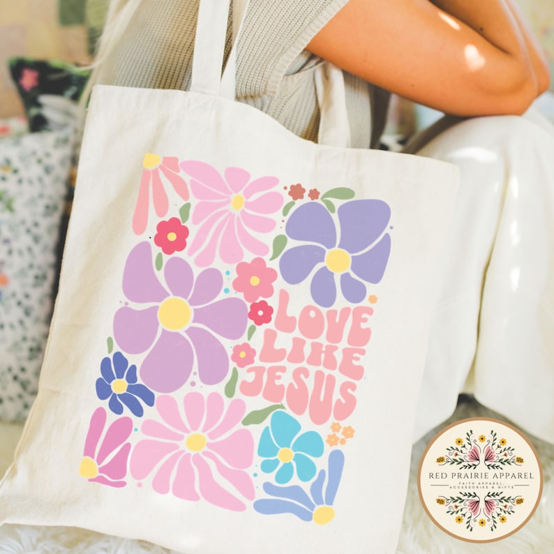 May include: A white canvas tote bag with a floral design and the text "Love Like Jesus" in a retro font. The flowers are pink, purple, blue, and yellow.
