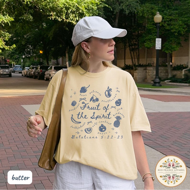 May include: Butter-colored t-shirt with a blue graphic of fruits and the words "Fruit of the Spirit" and "Galatians 5:22-23." The shirt is worn with a white cap and shorts. The brand logo is visible in the bottom right corner.