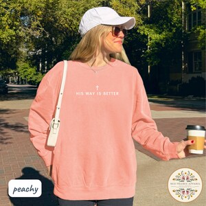 May include: Peach-colored sweatshirt with the text "HIS WAY IS BETTER" and a cross graphic. The sweatshirt is a crew neck style and has long sleeves. A white baseball cap and sunglasses are also visible.