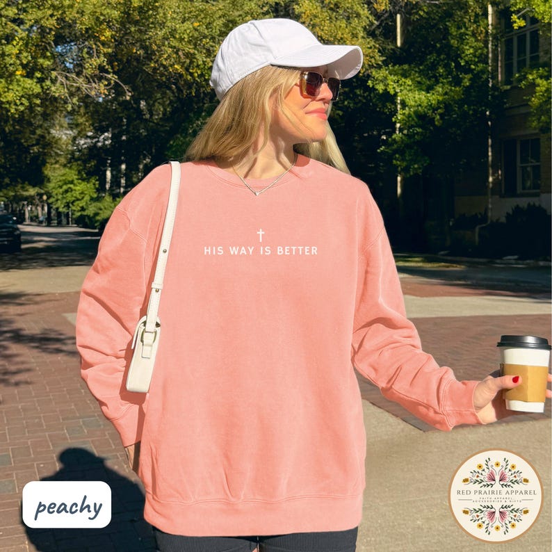 May include: Peach-colored sweatshirt with the text "HIS WAY IS BETTER" and a cross graphic. The sweatshirt is a crew neck style and has long sleeves. A white baseball cap and sunglasses are also visible.