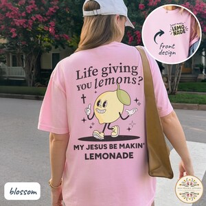 May include: A light pink t-shirt with the text "Life giving you lemons?" and "My Jesus be makin' lemonade." A cartoon lemon character is featured. The front design says "Jesus makin' lemonade."