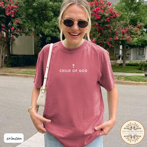 May include: Crimson-colored t-shirt with the text "CHILD OF GOD" and a cross symbol. The shirt is a short-sleeved crew neck style. The image also shows a white purse and sunglasses.