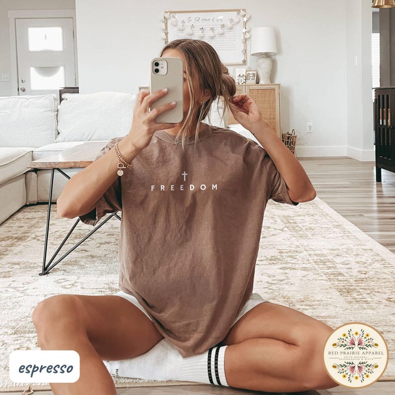 May include: Espresso-colored t-shirt with the word "FREEDOM" and a cross graphic. The shirt is a relaxed fit and is paired with white shorts and striped socks. The image is taken indoors.