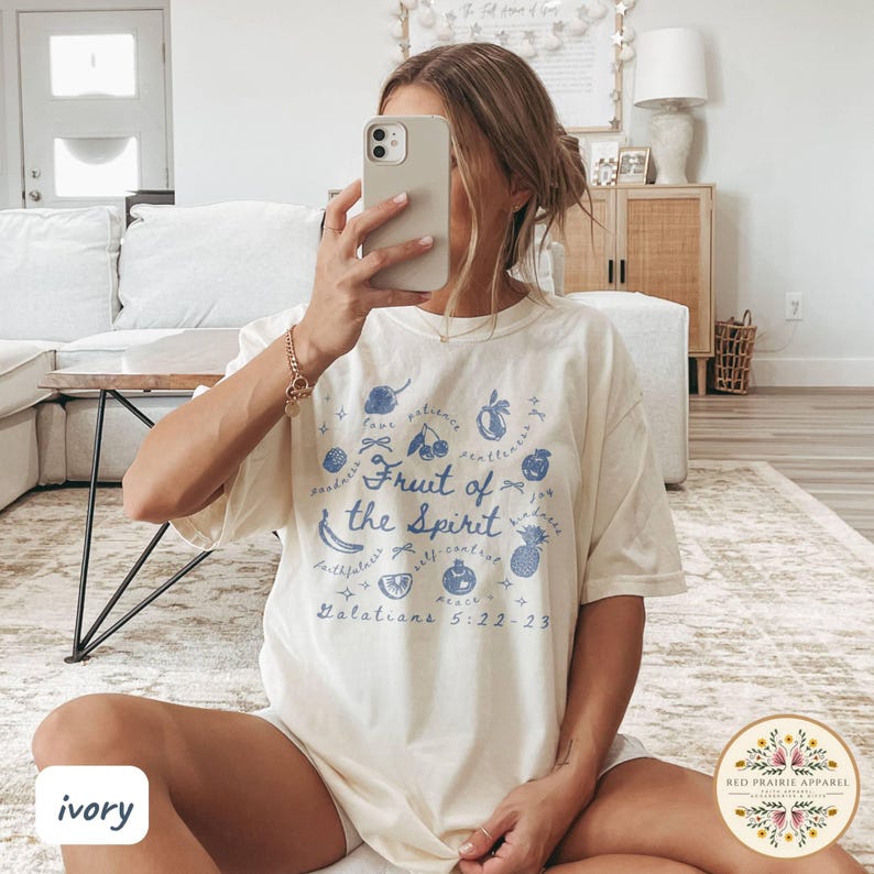 May include: Ivory-colored t-shirt with a blue graphic design. The design features the text "Fruit of the Spirit" surrounded by illustrations of fruits and the scripture reference Galatians 5:22-23. The shirt is from Red Prairie Apparel.