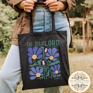 May include: Black tote bag featuring a floral design with blue flowers, green stems, and pink buds. The text "THE JOY of the LORD IS MY STRENGTH" is printed above the flowers. The bag is held by a person wearing a brown jacket and blue jeans.