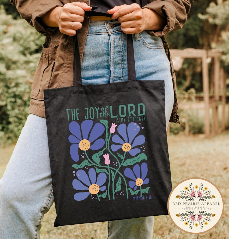 May include: Black tote bag featuring a floral design with blue flowers, green stems, and pink buds. The text "THE JOY of the LORD IS MY STRENGTH" is printed above the flowers. The bag is held by a person wearing a brown jacket and blue jeans.