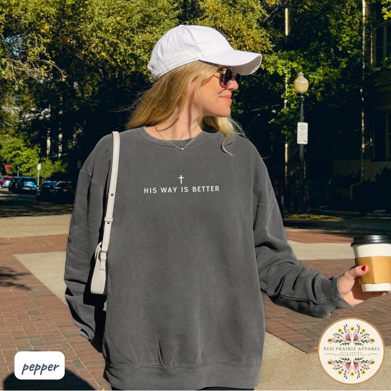 May include: A dark gray sweatshirt with the text "HIS WAY IS BETTER" and a cross graphic. The sweatshirt is worn with a white baseball cap and sunglasses. A coffee cup is held in the hand. The Red Prairie Apparel logo is visible.