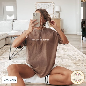May include: Espresso-colored t-shirt with the text "HIS WAY IS BETTER" and a cross graphic. The shirt is a relaxed fit and is paired with white shorts and striped socks. The image is taken indoors.