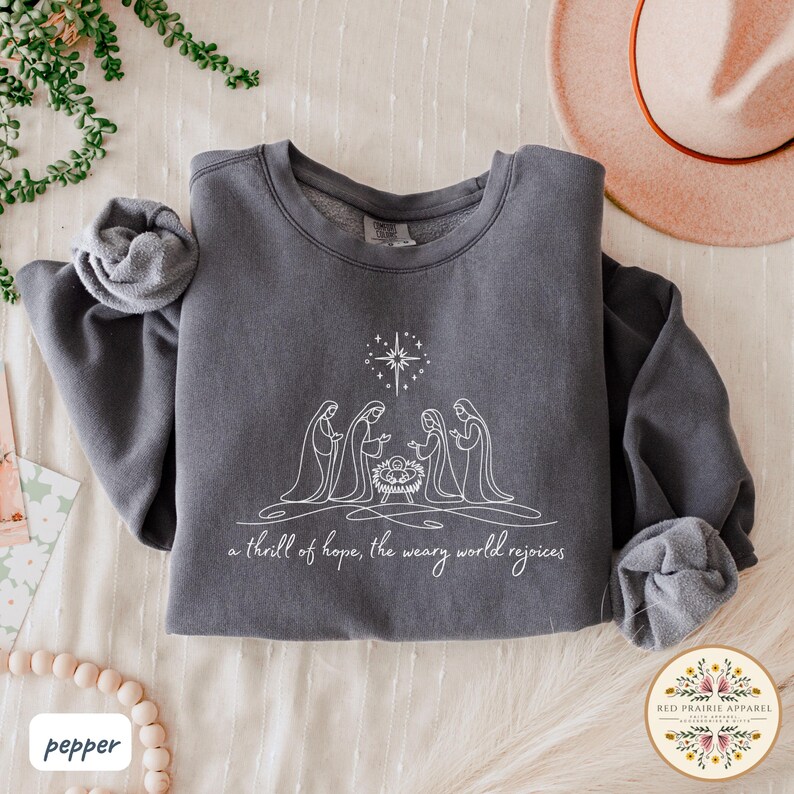 May include: A dark gray sweatshirt with a white line drawing of the nativity scene, including the star, Mary, Joseph, and baby Jesus. The text "a thrill of hope, the weary world rejoices" is below the image. The sleeves are rolled up.