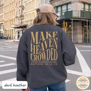 May include: A dark heather gray crewneck sweatshirt featuring gold text on the back that reads "MAKE HEAVEN CROWDED" in a wavy font, followed by the full text of Matthew 28:19-20. The garment is worn with a tan baseball cap in an outdoor setting.