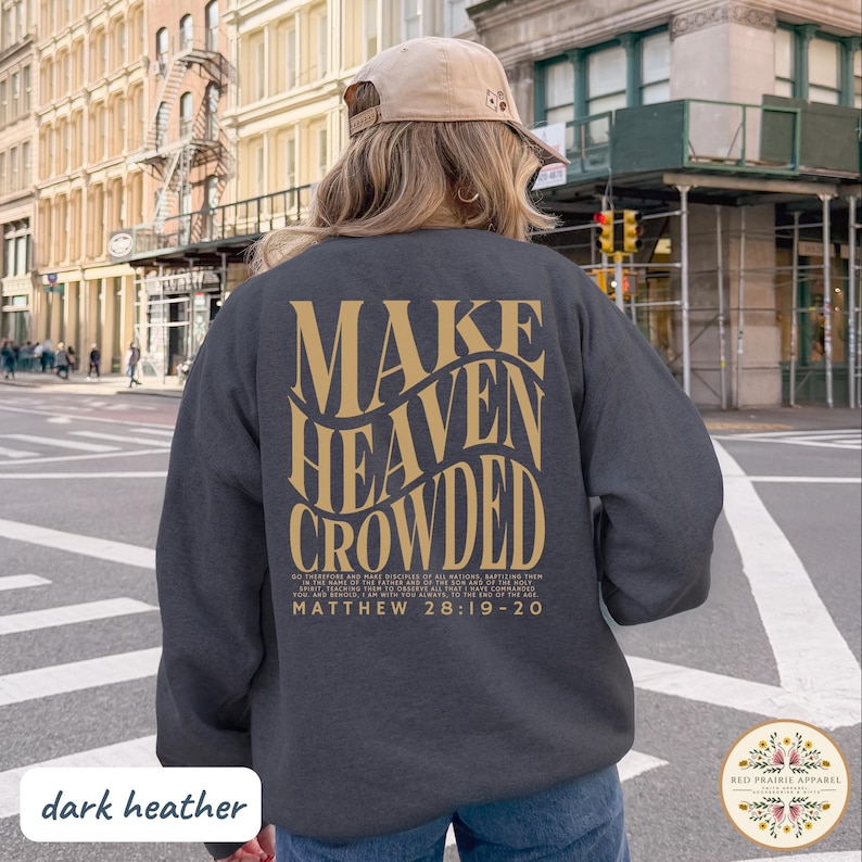 May include: A dark heather gray crewneck sweatshirt featuring gold text on the back that reads "MAKE HEAVEN CROWDED" in a wavy font, followed by the full text of Matthew 28:19-20. The garment is worn with a tan baseball cap in an outdoor setting.