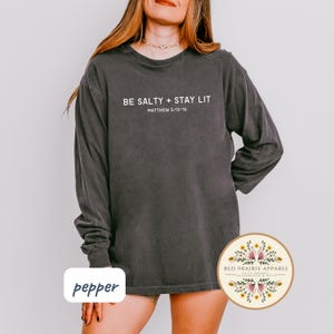 K&ouml;nnte beinhalten: Ein dunkelgraues Langarmshirt mit dem Aufdruck "BE SALTY + STAY LIT" und "MATTHEW 5:13-16" auf der Vorderseite. Das Shirt ist aus weichem, bequemem Material und hat eine lockere Passform.
