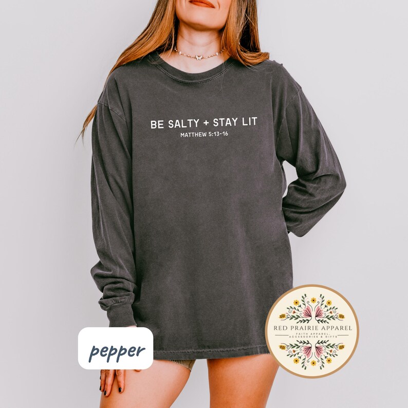 K&ouml;nnte beinhalten: Ein dunkelgraues Langarmshirt mit dem Aufdruck "BE SALTY + STAY LIT" und "MATTHEW 5:13-16" auf der Vorderseite. Das Shirt ist aus weichem, bequemem Material und hat eine lockere Passform.