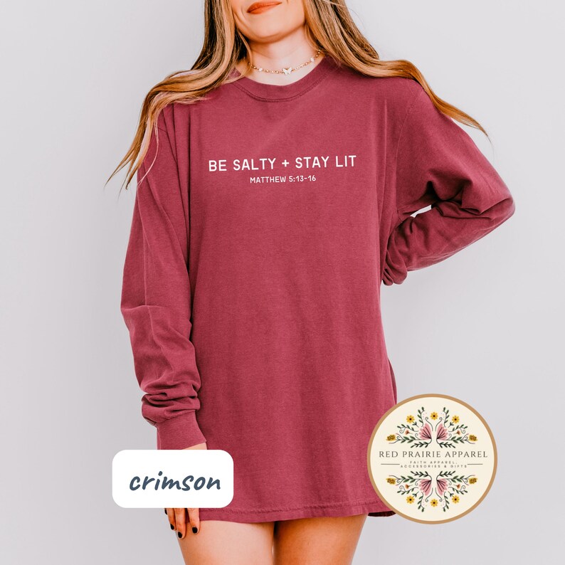 K&ouml;nnte beinhalten: Langarm-T-Shirt in der Farbe Crimson mit dem Text "BE SALTY + STAY LIT" und "MATTHEW 5:13-16" in Wei&szlig;. Das Shirt wird von einer Person mit langen braunen Haaren getragen.