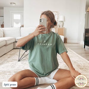May include: Light green t-shirt with the word "FREEDOM" and a cross graphic printed on the front. The shirt is a relaxed fit and is paired with white shorts and striped socks. The image is taken indoors.