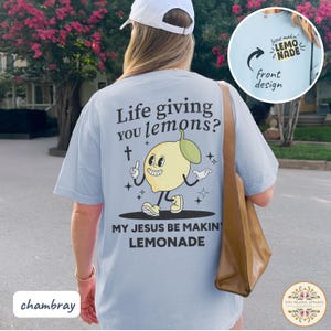 May include: Light blue chambray t-shirt with a cartoon lemon graphic and the text "Life giving you lemons? My Jesus be makin' lemonade." The front design shows the text "Jesus makin' lemonade."