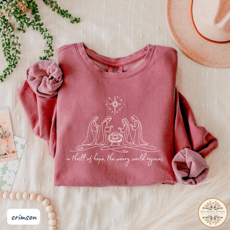 May include: Crimson-colored sweatshirt with a crew neck, featuring a white line drawing of the nativity scene. The text "a thrill of hope, the weary world rejoices" is below the image. The sweatshirt is folded on a light-colored surface.