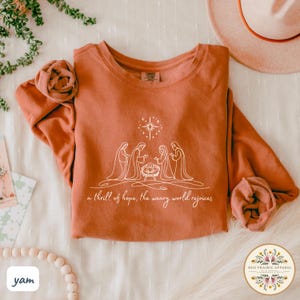 May include: Rust-colored sweatshirt with a white line drawing of the nativity scene, including the star of Bethlehem and the text "a thrill of hope, the weary world rejoices." The long-sleeved shirt is folded with a decorative flower.