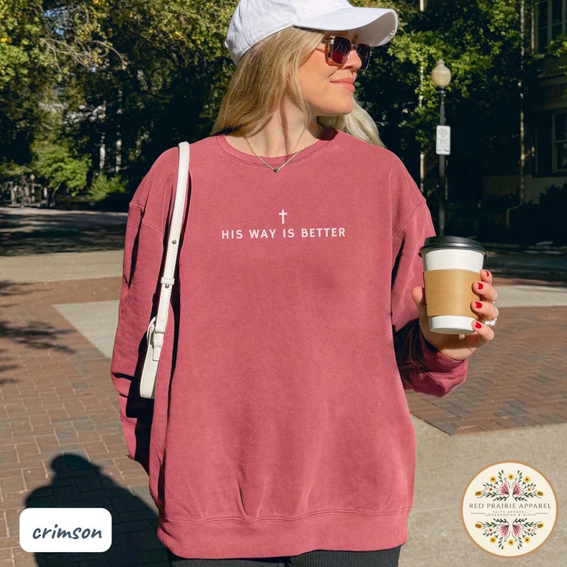 May include: Crimson-colored sweatshirt with the text "HIS WAY IS BETTER" and a cross graphic. The sweatshirt is a solid color and has a crew neck. The image also includes a white shoulder bag and a coffee cup.