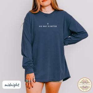 May include: A navy blue long-sleeve shirt with the text "HIS WAY IS BETTER" and a cross symbol in white. The shirt is a relaxed fit and has a crew neck. The word "midnight" is in the lower left corner.