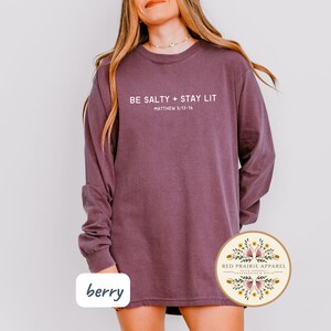 K&ouml;nnte beinhalten: Ein lang&auml;rmeliges, beerfarbenes Shirt mit dem Text "BE SALTY + STAY LIT" und "MATTHEW 5:13-16" in Wei&szlig;. Das Shirt hat einen Rundhalsausschnitt und ist aus einem weichen, bequemen Stoff gefertigt.