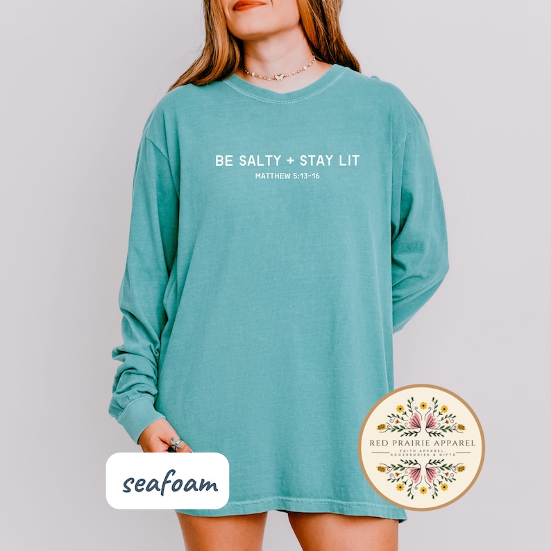 K&ouml;nnte beinhalten: Langarm-T-Shirt in Seefraben mit dem Text "BE SALTY + STAY LIT" und "MATTHEW 5:13-16" in Wei&szlig;. Das Shirt hat eine lockere Passform und einen Rundhalsausschnitt.