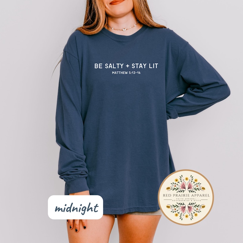 K&ouml;nnte beinhalten: Ein marineblaues Langarmshirt mit dem Text "BE SALTY + STAY LIT" und "MATTHEW 5:13-16" in Wei&szlig;. Das Shirt wird von einem Model getragen und der Text "midnight" ist in der linken unteren Ecke des Bildes sichtbar.