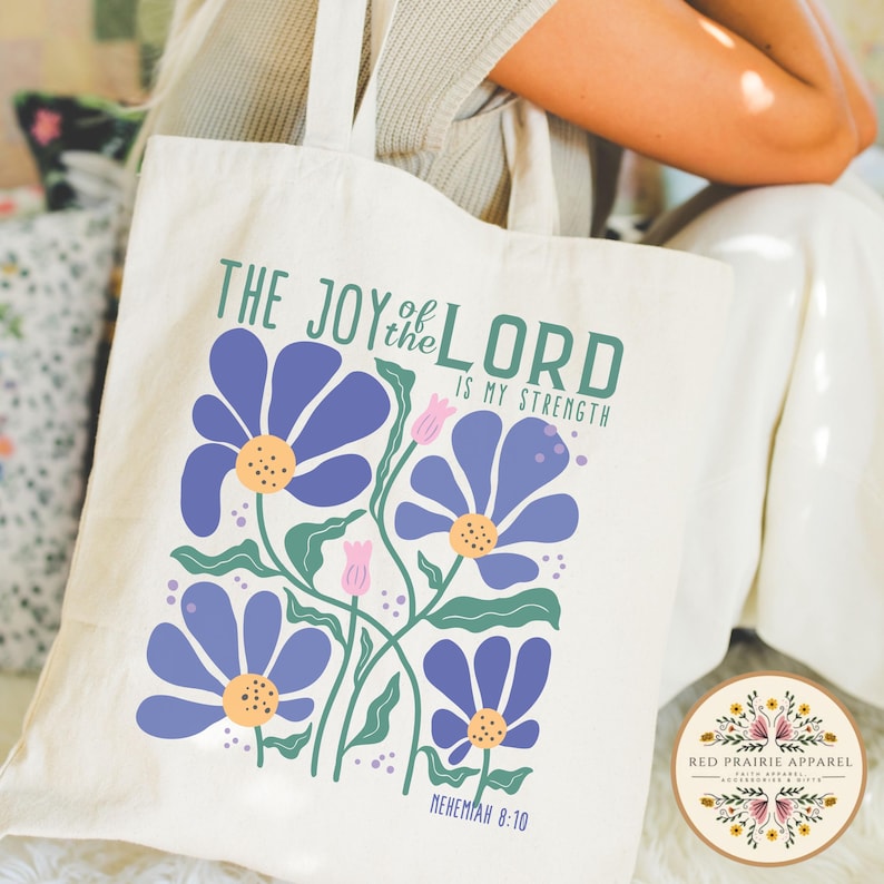 May include: Cream-colored tote bag featuring a floral design with blue flowers, green stems, and yellow centers. The text "THE JOY of the LORD IS MY STRENGTH" is printed above the flowers, with "NEHEMIAH 8:10" below. A circular logo is in the bottom right corner.