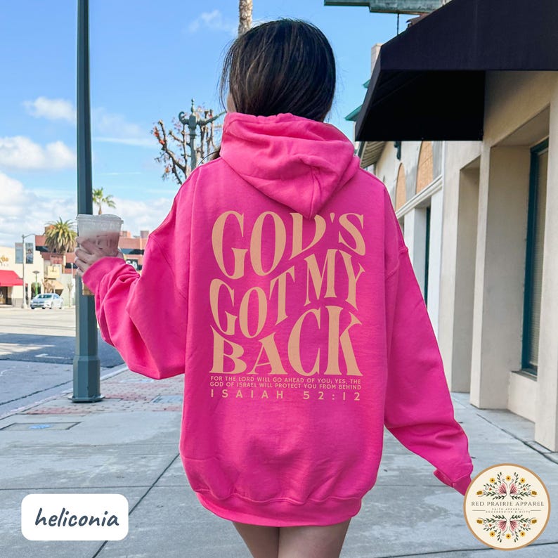 May include: A bright pink hoodie with the text "GOD'S GOT MY BACK" in a tan color on the back. The hoodie also features the Bible verse Isaiah 52:12. The brand name "Red Prairie Apparel" is visible on the bottom right.