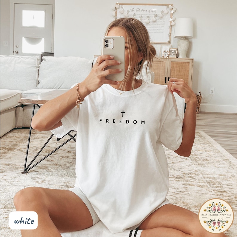 May include: White t-shirt with a black cross and the word "FREEDOM" printed on the front. The shirt is a casual style, suitable for everyday wear. The image also shows a logo in the bottom right corner.