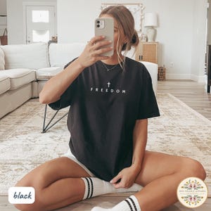 May include: Black t-shirt with the word "FREEDOM" and a cross graphic in white. The shirt is worn with white shorts and striped socks. The image is taken indoors.