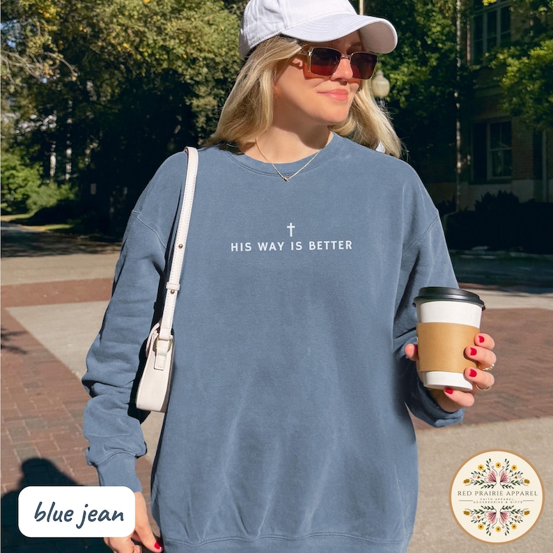 May include: Blue jean colored sweatshirt with the text "HIS WAY IS BETTER" and a cross graphic. The sweatshirt is a solid color and has long sleeves. The image also includes a white purse, sunglasses, and a coffee cup.