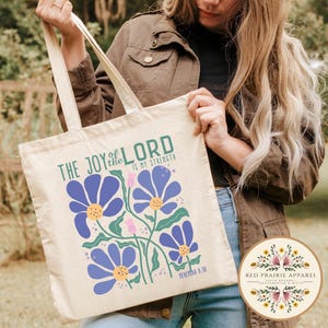 May include: Cream-colored tote bag featuring a floral design with blue flowers, green stems, and pink buds. The text "THE JOY of the LORD IS MY STRENGTH" is printed in green. A round logo with floral accents is visible.