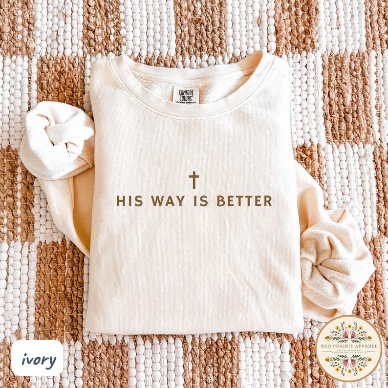 May include: Ivory-colored sweatshirt with the text "HIS WAY IS BETTER" and a cross graphic. The long-sleeved top is made by Comfort Colors and has a crew neck. The sweatshirt is displayed on a textured, neutral-toned surface.