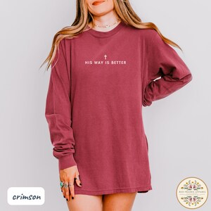May include: Crimson long-sleeve shirt with the text "HIS WAY IS BETTER" and a cross symbol in white. The shirt is a solid crimson color and has a relaxed fit. The shirt is from Red Prairie Apparel.