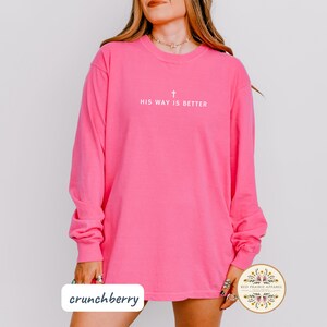 May include: A long-sleeved, bright pink shirt with the text "HIS WAY IS BETTER" and a cross symbol printed in white. The shirt is a solid color and has a casual style. The word "crunchberry" is printed on the bottom left.