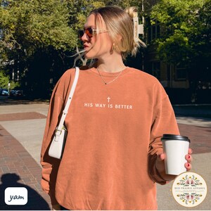 May include: A rust-colored sweatshirt with the text "HIS WAY IS BETTER" and a cross graphic. The long-sleeved top is worn by a person outdoors, paired with a white purse. A coffee cup is held in the hand.
