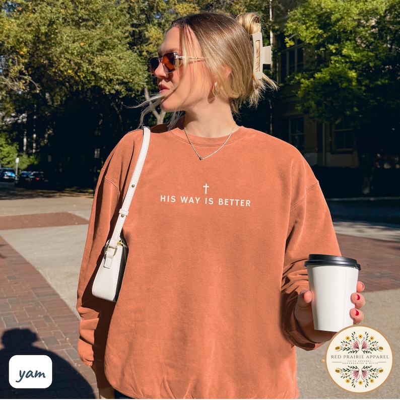 May include: A rust-colored sweatshirt with the text "HIS WAY IS BETTER" and a cross graphic. The long-sleeved top is worn by a person outdoors, paired with a white purse. A coffee cup is held in the hand.