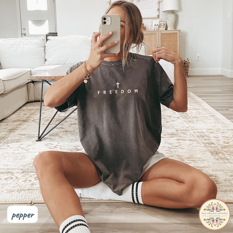 May include: Dark gray t-shirt with the word "FREEDOM" and a cross printed in white. The shirt is a crew neck style and has short sleeves. The model is wearing white shorts and white socks with black stripes.