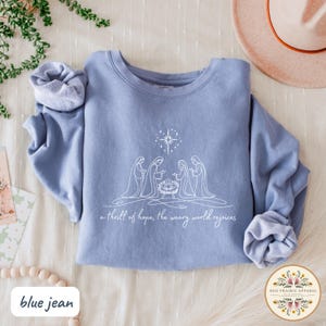 May include: A blue jean-colored sweatshirt featuring a white line drawing of the nativity scene with a star above. The text "a thrill of hope, the weary world rejoices" is below the image. The Red Prairie Apparel logo is in the bottom right.