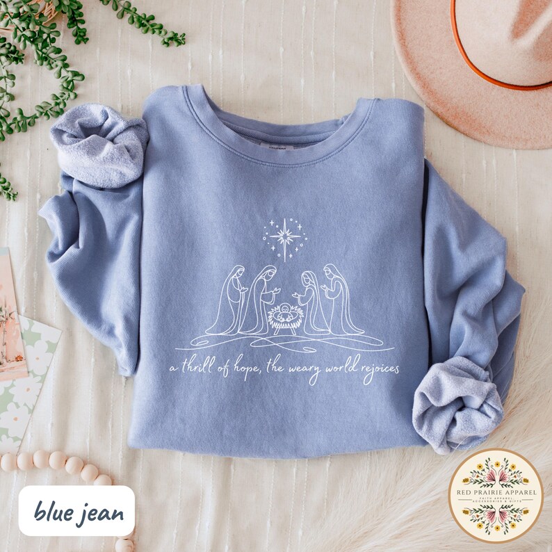 May include: A blue jean-colored sweatshirt featuring a white line drawing of the nativity scene with a star above. The text "a thrill of hope, the weary world rejoices" is below the image. The Red Prairie Apparel logo is in the bottom right.