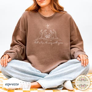 May include: Espresso-colored sweatshirt featuring a white line drawing of the nativity scene with a star above. The text "a thrill of hope, the weary world rejoices" is below the image. The sweatshirt is a relaxed fit.
