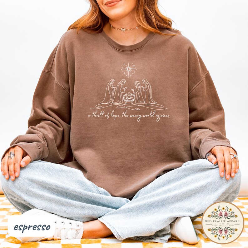 May include: Espresso-colored sweatshirt featuring a white line drawing of the nativity scene with a star above. The text "a thrill of hope, the weary world rejoices" is below the image. The sweatshirt is a relaxed fit.