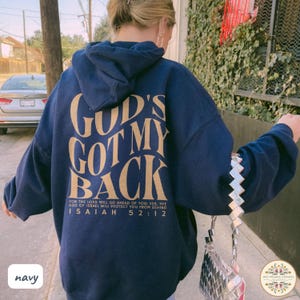 May include: Navy blue hoodie with the text "GOD'S GOT MY BACK" in a gold color on the back. The hoodie also features the Bible verse Isaiah 52:12. The hoodie is a comfortable, casual style.
