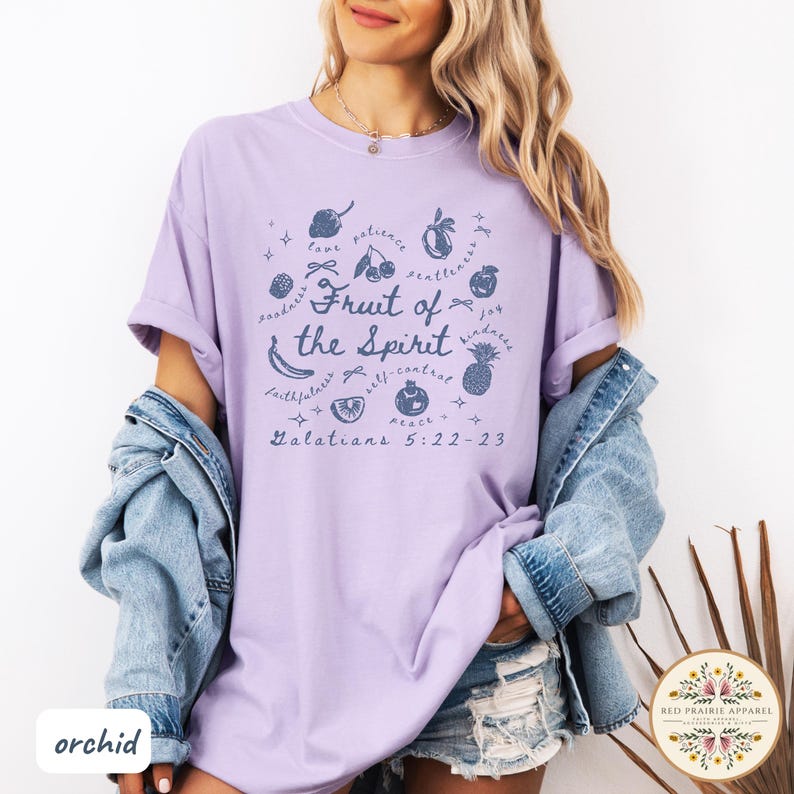 May include: A light purple t-shirt with the text "Fruit of the Spirit" and related words and illustrations. The text "Galatians 5:22-23" is also printed on the shirt. The shirt is part of the Red Prairie Apparel collection.